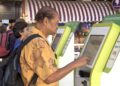 Homem idoso com camisa florida amarela usa totem digital de autoatendimento no aeroporto acompanhado por outros passageiros