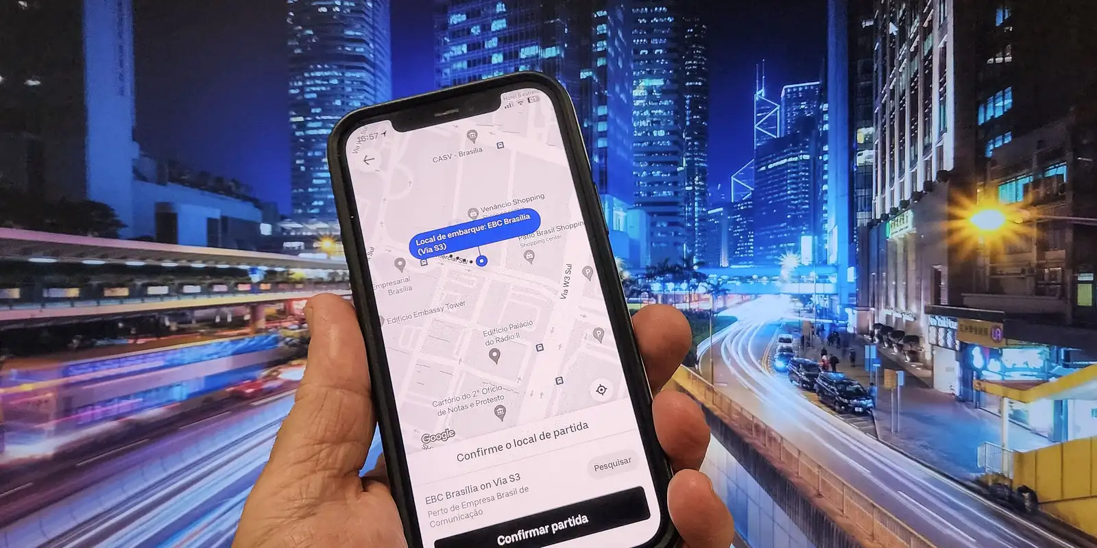 Adeus tarifa de cadastro: CMN determina que bancos não podem cobrar taxa de motoristas no Move Aplicativos 2 Mão segura celular exibindo mapa com local EBC Brasília sobre fundo de cidade iluminada à noite