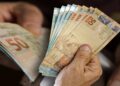 Mão de pessoa idosa segura várias cédulas de cinquenta reais, foco nas cédulas do Banco Central do Brasil