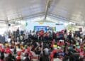 Autoridades no palco com público diverso e painel "Governo do Brasil na Rua" em evento coberto