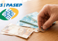 Mão pegando moedas ao lado de notas de real com logo do PIS/PASEP na mesa.