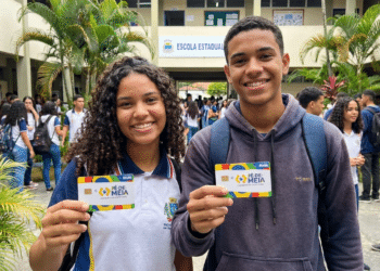 Dois estudantes uniformizados sorrindo e mostrando cartões do programa Pé-de-Meia na frente de escola estadual