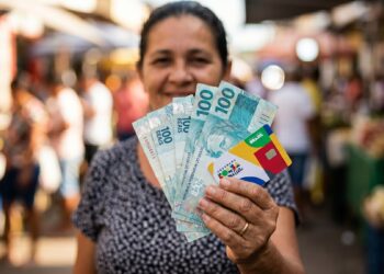 Mulher sorrindo segura notas de 100 reais e cartão do Bolsa Família em feira a céu aberto