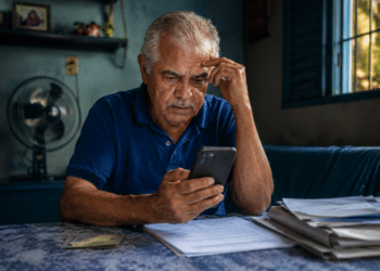 Idoso analisa celular com expressão preocupada à mesa com documentos ao lado em casa