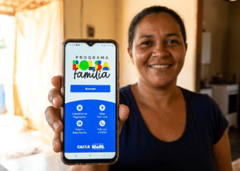 Mulher sorridente de camiseta azul marinho mostra celular com app Bolsa Família, logotipo Caixa e Brasil visíveis