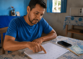 Homem aponta para data em calendário de mesa ao lado de celular e notas de real