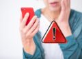 Mulher preocupada olha o celular ao lado de ícone vermelho de aviso