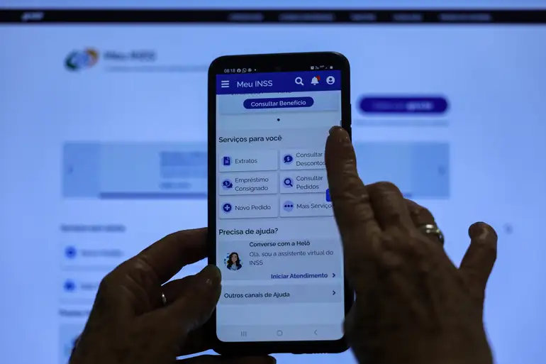 Governo exige 8 documentos atualizados de idosos: quem não regularizar pode ter benefícios suspensos 2 Mãos acessando aplicativo Meu INSS no celular com tela de computador ao fundo