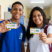 Dois jovens estudantes uniformizados mostram cartões do programa Pé-de-Meia em corredor de escola